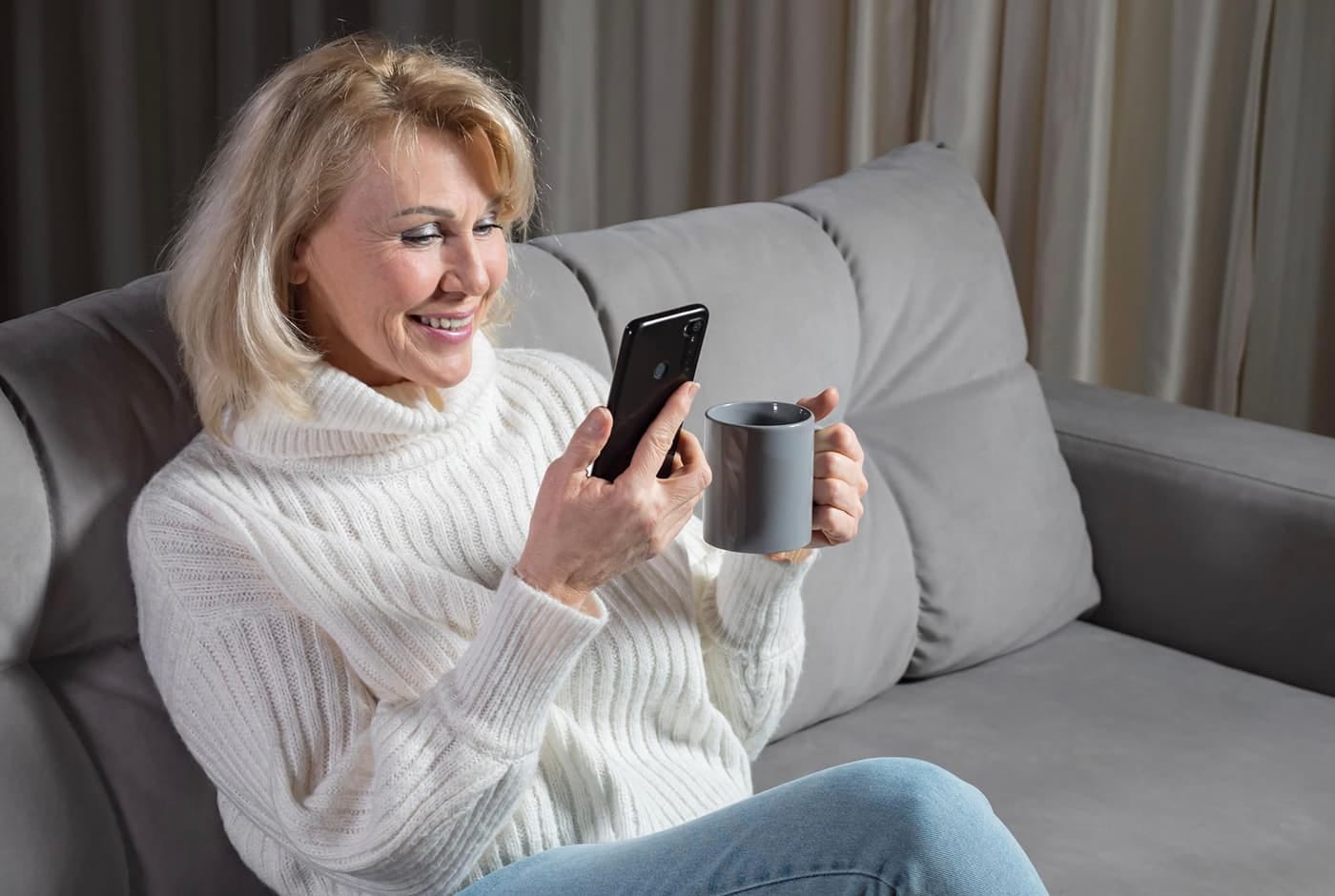 Mature blond woman in a white sweater enjoying online conversation sitting on a comfortable sofa with a cup of well-brewed coffee